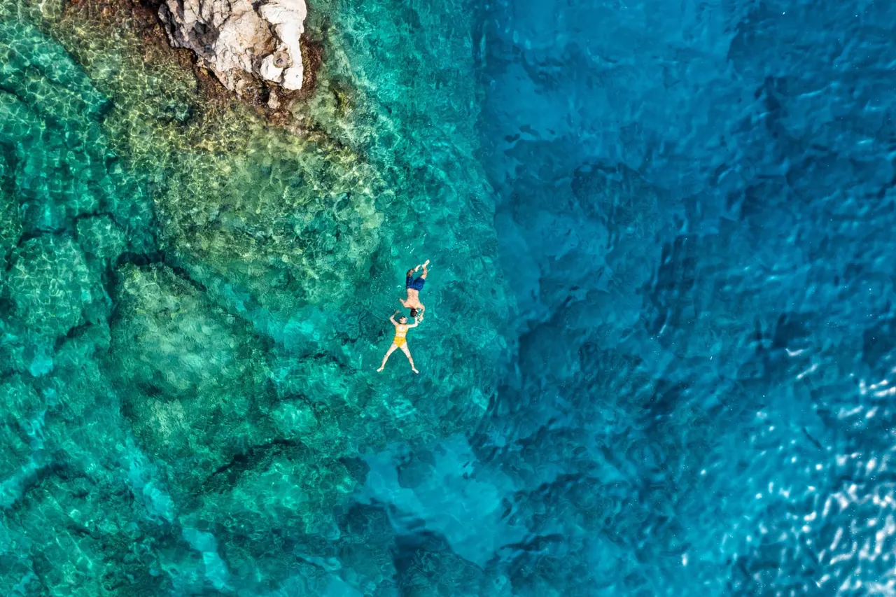 Aerial view of two people floating in clear turquoise water beside rocks