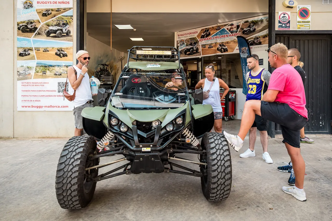 Group of people standing around a road buggy while one person sits at the wheel