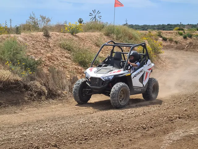 Child with helmet driving a white kids buggy on a dirt track, flowering shrubs and a windmill behind