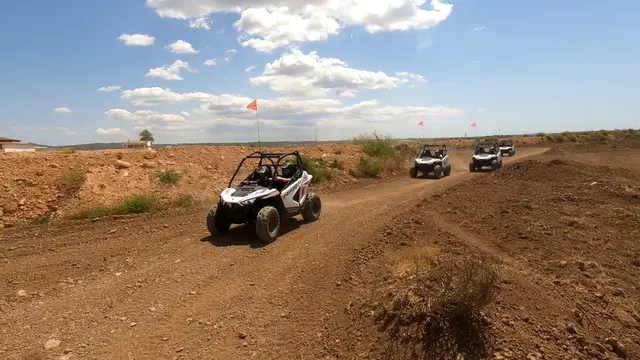 Several white kids buggies driving in a line on the dirt circuit, two helmeted people in the front buggy