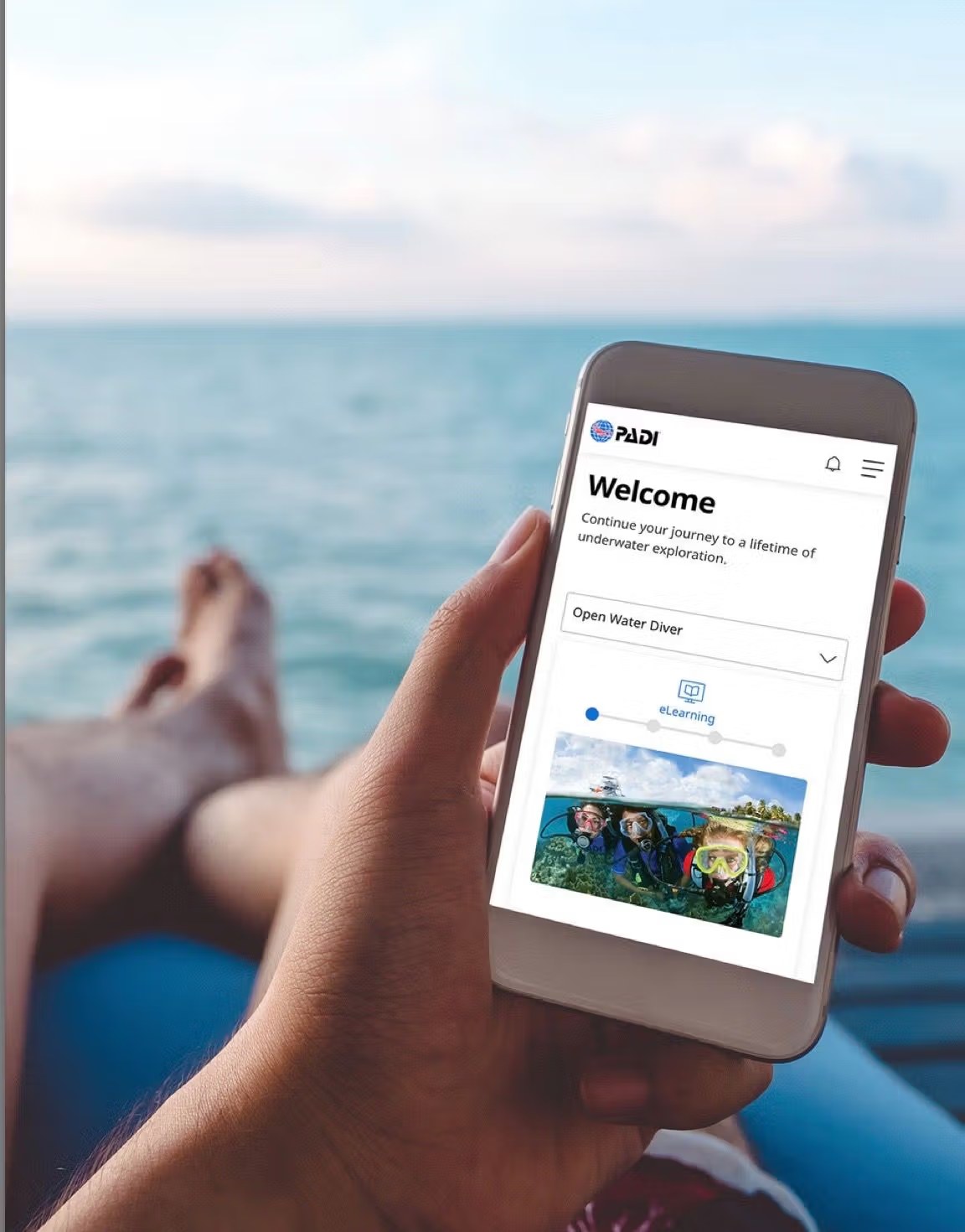 A hand holding a smartphone showing the PADI Open Water Diver eLearning course, with the sea in the background
